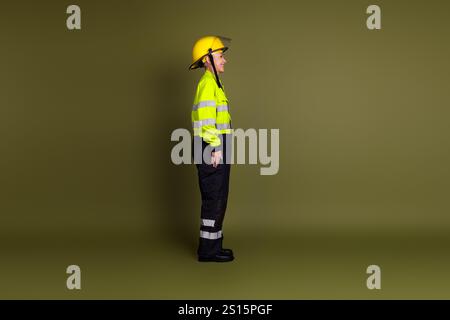 Mature woman in safety gear standing confidently against a khaki backdrop, showcasing resilience in a high-visibility uniform and hardhat Stock Photo