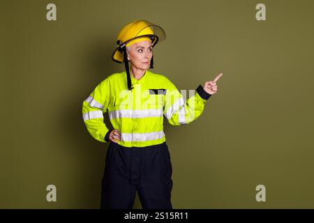 Mature female professional in high visibility workwear points decisively, standing against a khaki background Stock Photo