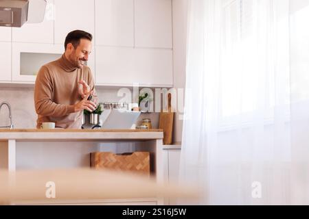 Confident male freelancer gesturing and explaining over video call on laptop while working in kitchen Stock Photo
