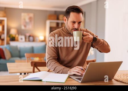 Portrait of handsome programmer drinking coffee and using laptop while working at desk in home office Stock Photo