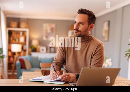 Serious businessman looking away thoughtfully while writing notes and working over laptop at desk Stock Photo
