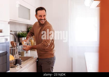 Handsome man smiling and looking away while cleaning drinking glass with sponge in kitchen sink Stock Photo