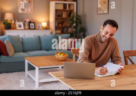 Portrait of handsome mid adult man smiling and writing in diary while using laptop on desk in living room Stock Photo