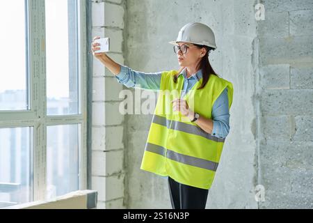Female industrial worker in protective vest helmet using smartphone at construction site Stock Photo