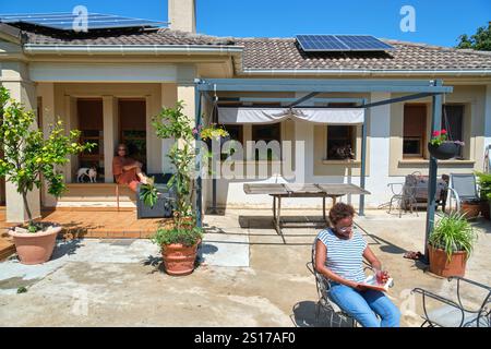 Two women are relaxing in a sunny backyard; one is sitting on the porch with a dog, while the other reads a book while holding a drink. The backyard f Stock Photo
