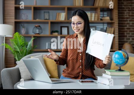 Asian woman conducting online lesson from home. She sits on sofa with laptop, holding a whiteboard marked Lesson, engaged in interactive teaching session, conveying education and technology. Stock Photo