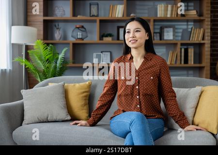 Asian woman relaxing on sofa wearing casual polka dot shirt in modern home. Surrounded by stylish decor, she appears calm and content, embodying a sense of comfort and tranquility. Stock Photo