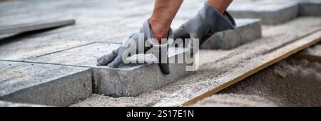A worker wearing gloves carefully places paving stones on a sand base, ensuring an even surface for a durable and smooth pathway. Wide view image. Stock Photo