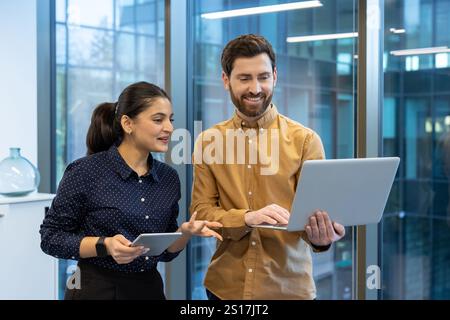 Two colleagues man and woman inside office standing by window talking, group of office workers using laptop discussing new work project. Stock Photo