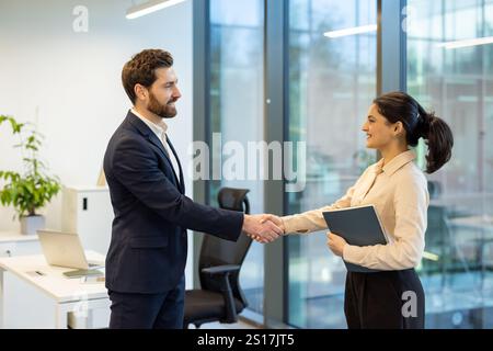 Two business colleagues shaking hands, businesswoman and businessman smiling happily greeting each other, colleagues working inside office. Stock Photo