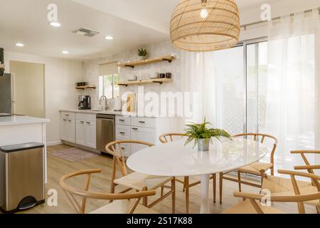 Bright, modern kitchen and dining area with white cabinets, light wood dining chairs, a white oval table, and a large woven pendant light. Stock Photo
