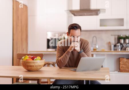 Focused male entrepreneur with hands clasped analyzing data over laptop on dining table at home Stock Photo
