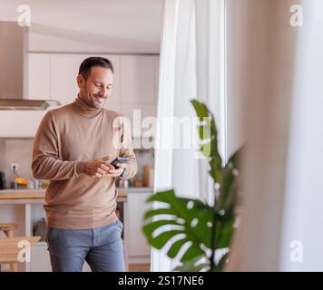 Mid adult handsome man checking social media apps over phone while standing by window in home office Stock Photo
