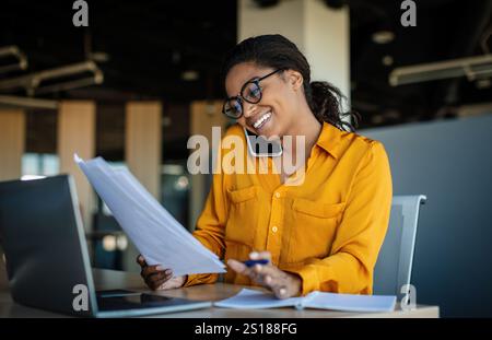 Corporate communication. Happy black female manager working with papers and talking on mobile phone Stock Photo