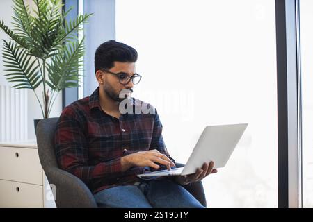 Young Indian male in casual attire using laptop seated by large window. Bright natural light fills modern workspace, creating an inspiring environment for remote work. Stock Photo