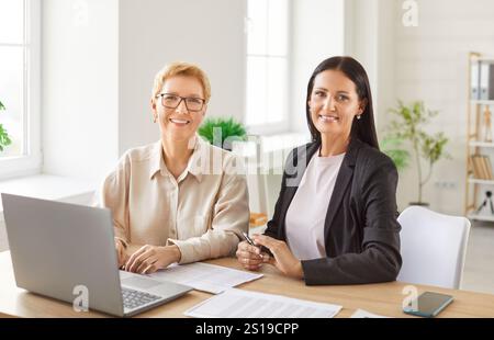 Female coworkers working in office sitting at desk with laptop and looking cheerful at camera Stock Photo