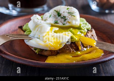 Cutting poached egg with runny egg yolk over bread toast with mashed avocado spread in plate, close up. Healthy breakfast or lunch food Stock Photo