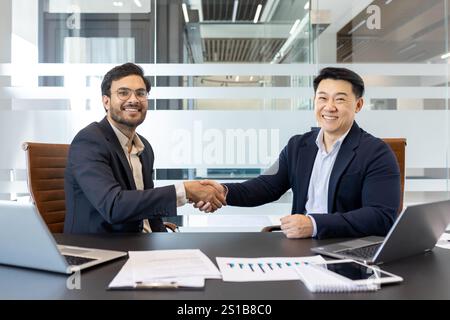 Handshake, two businessmen inside the office, business meeting agreeing on a contract, men smiling happily and looking at the camera, working with a laptop in a meeting room inside the office. Stock Photo