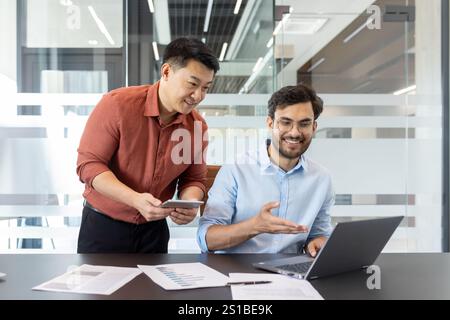 Two diverse colleagues discussing new project startup, male business team at workplace in casual clothes working with laptop and tablet computer, discussing new software. Stock Photo