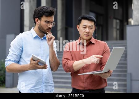 Two office workers discussing a work project outside an office building, miscellaneous business meeting of office company workers, discussing looking at a laptop, serious and focused. Stock Photo