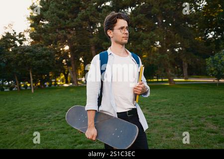 Young man with skateboard and books standing in park during sunset Stock Photo