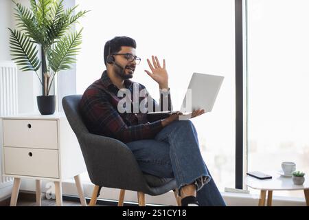 Indian young man sitting in chair having video call on laptop. Man wearing headset and plaid shirt, appears engaged and happy. Working from home concept. Stock Photo