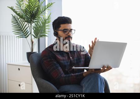 Young Indian man working remotely with laptop and headset, sitting comfortably. Engaged in online communication, focused on professional tasks. Stock Photo
