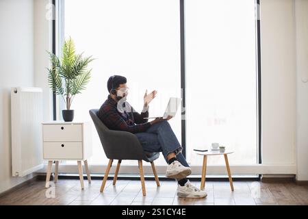 Indian man wearing headphones works remotely on laptop in home setting. Casual attire suggests comfort as he communicates. Natural light and plants add to relaxed ambiance. Stock Photo