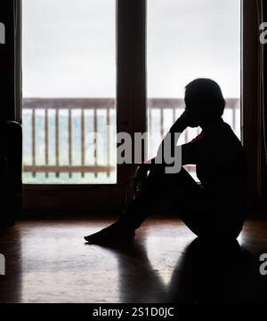 Lonely woman sitting on the floor by the glass door, looking outside. Stock Photo