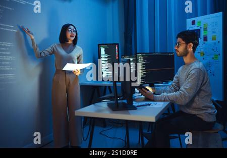 asian woman developers collaborating on a coding project, with one member presentation at code projected on the wall while the other works on multiple Stock Photo