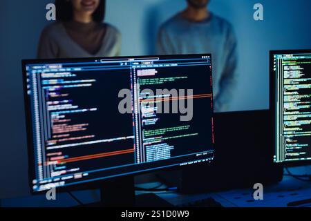 Close-up of computer screens displaying code, with developers collaborating in the background Stock Photo