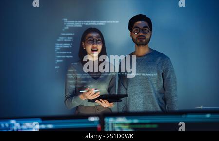Asian Two software developers engaged in collaborative coding session, with code projected onto the wall and multiple computer screens displaying Stock Photo