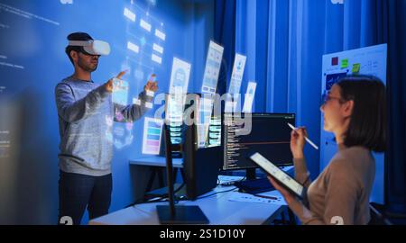 developer uses a VR headset to interact with projected interface designs, while colleagues discuss and code on screens at office Stock Photo