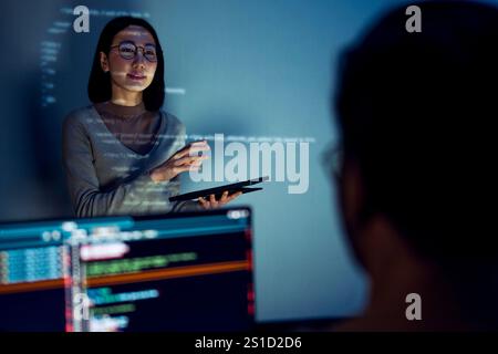 asian woman developers collaborating on a coding project, with one member presentation at code projected on the wall while the other works on multiple Stock Photo