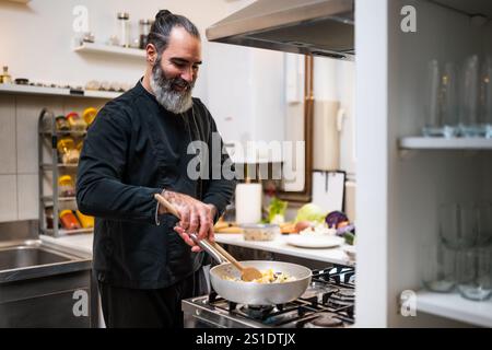 Professional chef is preparing meal in restaurant's kitchen. He makes a vegetarian lunch. He is mixing food in cooking pan. Stock Photo