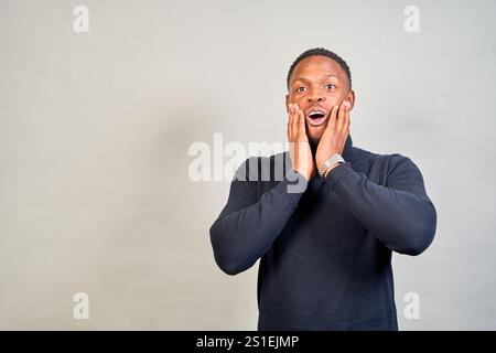 Surprised young man in a cozy sweater holds his face, showing wonder and joy against a gray background. His african american features add uniqueness t Stock Photo