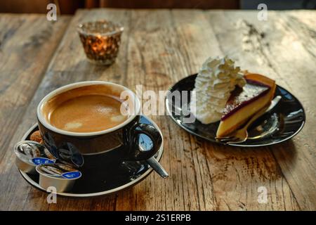 Close-up of a cup of coffee served with a slice of cake topped with whipped cream, set on a rustic wooden table with warm ambient lighting. Stock Photo