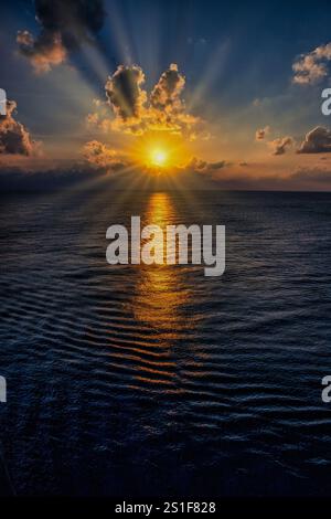 Golden sunlight breaks through dramatic clouds, casting shimmering reflections across tranquil ocean waves at sunset Stock Photo