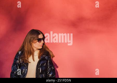Woman with sunglasses leaning against a vibrant red wall on a sunny day in an urban area Stock Photo