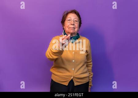 Elderly professor with glasses and mustard-colored cardigan making come here gesture with hand on purple background, inviting students to approach Stock Photo