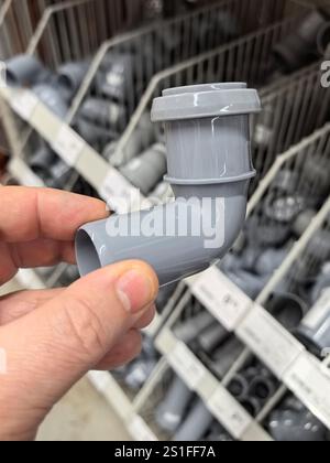 A hand holds a gray plumbing pipe elbow while browsing through various plumbing supplies in a well-stocked hardware store aisle. The organized shelves Stock Photo