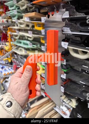 A person holds a uniquely designed tool featuring a bright orange handle in a vibrant hardware store aisle, filled with various tools and materials. Stock Photo