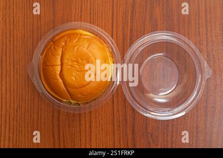 A single freshly made butter bun displayed inside a transparent plastic container on a wooden table. Perfect for presentations of fast food. Stock Photo