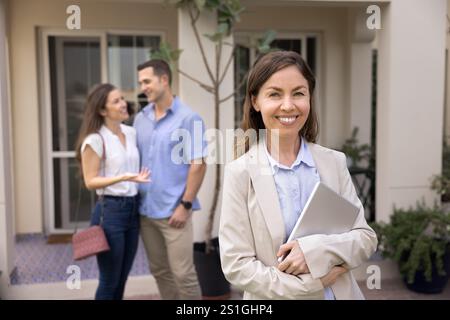 Happy realtor posing outdoors with accommodation and renters in background Stock Photo