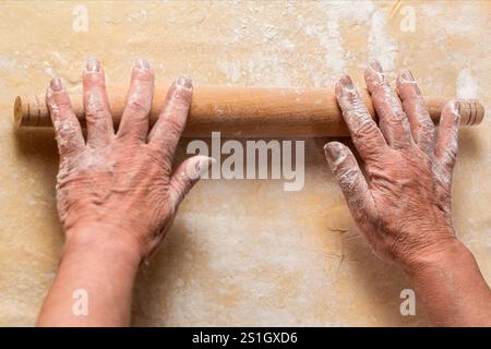 Closeup of man rolling dough with rolling pin, cooking process top view. Woman old hands rolling out dough in flour with rolling pin in bakery. Female Stock Photo