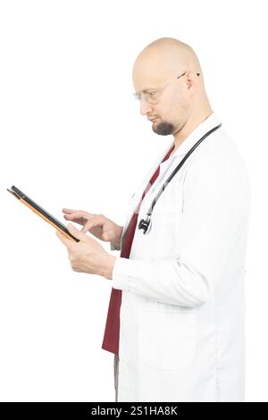 A middle-aged male doctor with light skin, glasses, and a stethoscope around his neck is wearing a white lab coat. He is attentively using a tablet, l Stock Photo