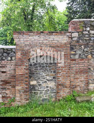 A weathered wall combines stone and brick materials, featuring an arched niche. Lush greenery surrounds the structure, highlighting its historical charm Stock Photo