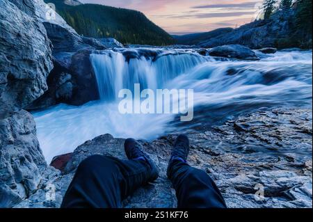 POV legs of traveler sitting and relaxing beside rapids waterfall flowing in the forest during evening at Elbow falls, Kananaskis, Alberta, Canada Stock Photo