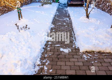 Brick garden pathway cleared of snow, leading to parked car, surrounded by snow-covered trees and shrubs. Stock Photo