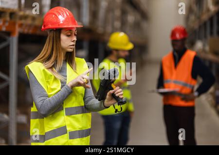 Warehouse employees wearing safety vests and helmets utilize technology for inventory management. The team collaborates in a large industrial setting, Stock Photo